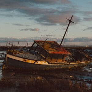 A weathered fishing boat, leaning at an angle in shallow water, with a rusty red roof and overgrown moss, set against a cloudy sky and distant silhouettes of other boats and land.