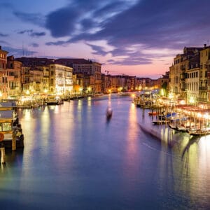 A serene evening view of the Grand Canal in Venice, with illuminated buildings lining the waterway, boats docked at piers, and a vibrant sky transitioning to twilight.