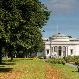 A historical building with a dome and columns is visible behind lush green trees in a park, with benches and flower beds in the foreground.