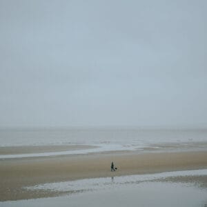 A solitary figure walks a dog along a desolate beach under overcast skies, with patches of wet sand reflecting the muted light.
