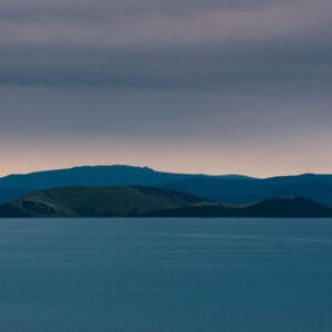 A distant view of two islands on a calm body of water under a cloudy sky, with mountains visible in the background.