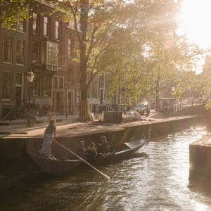 A serene canal scene in Amsterdam at sunset, featuring a small rowing boat with four people, surrounded by historic buildings and trees. The soft sunlight creates a warm glow over the water, and bicycles are parked along the canal side.