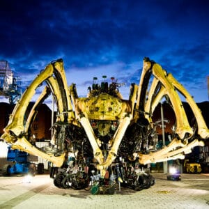 A large mechanical spider sculpture with elongated limbs is positioned in an outdoor area during twilight, with a dark blue sky and scattered clouds. Surrounding machinery and vehicles are visible, along with streetlights illuminating the scene.