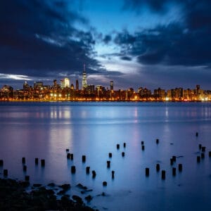 A twilight view of a city skyline reflecting in water, with silhouetted wooden posts in the foreground and bright city lights lining the horizon against a dramatic sky.