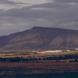 A panoramic view of rolling hills and distant mountains under a cloudy sky, with fields and a few scattered buildings in the foreground.