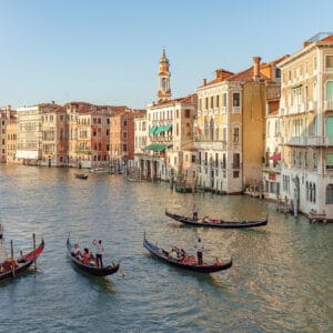A scenic view of a canal in Venice, featuring several gondolas with passengers and gondoliers, surrounded by historic buildings with balconies and shutters, under a clear blue sky.
