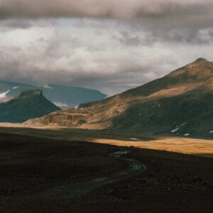 A winding dirt road leads through a vast, rugged landscape with mountains under a cloudy sky, showcasing a mix of shadows and light across the terrain. Snow patches are visible on the higher peaks.