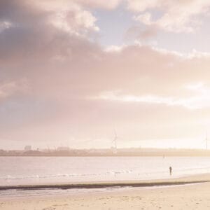 A tranquil beach scene with a lone figure walking along the shoreline, against a backdrop of soft clouds and gentle waves. Wind turbines are visible in the distance along with a hazy skyline.