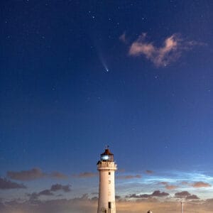 A lighthouse stands on the shore under a starry night sky, with a comet visible in the background. The scene is enhanced by clouds and distant city lights.