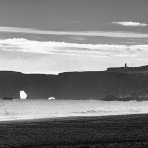 A lone figure walking along a sandy beach, with a large rock formation featuring an arch in the background, and a lighthouse visible on a distant cliff under a cloudy sky.