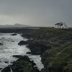 A coastal scene featuring a white house situated on rocky cliffs overlooking turbulent waves, with a grey, overcast sky and distant hills in the background.