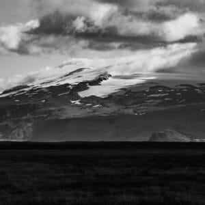 A dramatic black-and-white landscape of a snow-capped mountain with a glacier, partially obscured by clouds, set against a dark, textured foreground.