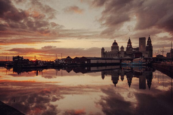 A scenic view of a marina at sunset, featuring silhouetted boats and buildings reflecting in the water, with cloudy skies and a warm orange glow.