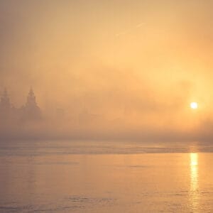 A foggy sunrise over a body of water, with silhouetted buildings and towers in the background, casting soft golden hues in the mist.