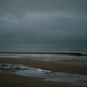 A cloudy beach scene with a vast, wet sandy shore and a distant breakwater. A solitary figure stands near the water's edge, while patches of wet sand reflect the overcast sky.