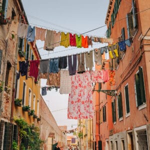 A narrow alleyway lined with colourful buildings, featuring laundry hanging between them on clotheslines, with various garments visible. There are potted plants on window sills and a clear sky overhead.
