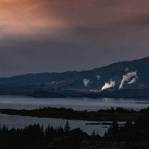 A scenic view of a lake surrounded by mountains, with steam rising from geothermal plants in the background under a moody sky.