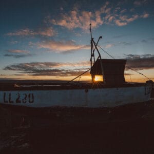 A silhouette of a fishing boat with the number "LL230" on its side, against a sunset sky featuring clouds, with sunlight shining through the boat's cabin.
