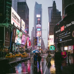 A rainy day in Times Square, New York City, featuring bright digital billboards and advertisements. Two people are walking with an umbrella, while yellow taxis are parked nearby. The wet pavement reflects the vibrant lights of the surroundings.