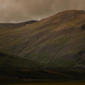 A rugged mountain landscape with sloping, textured hills in varying shades of green and brown under a cloudy sky.