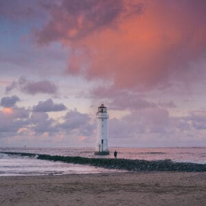 A lighthouse stands on a rocky jetty extending into the sea at sunset, surrounded by clouds illuminated in shades of pink and purple. A solitary figure is seen on the jetty near the lighthouse.