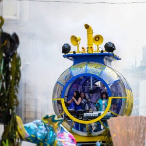 A vibrant blue and yellow music-themed vehicle resembling a submarine, with three musicians visible inside, performing during a parade surrounded by smoke and festive decorations.