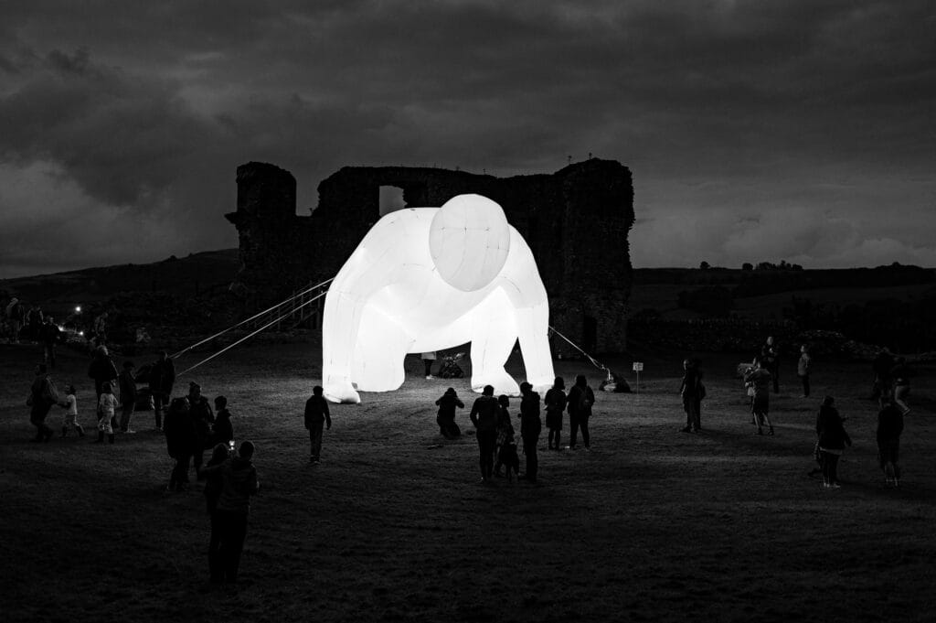 A large, illuminated white male sculpture is displayed outdoors at dusk, surrounded by a crowd of people, with a historic stone ruin in the background and a cloudy sky above, creating a dramatic atmosphere.