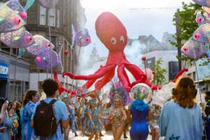 A vibrant carnival scene in Liverpool featuring a large, animated octopus puppet towering above participants, who are dressed in colorful costumes with fish-themed elements and sequins, surrounded by whimsical fish puppets on sticks, with a festive atmosphere and smoke effects in the background.