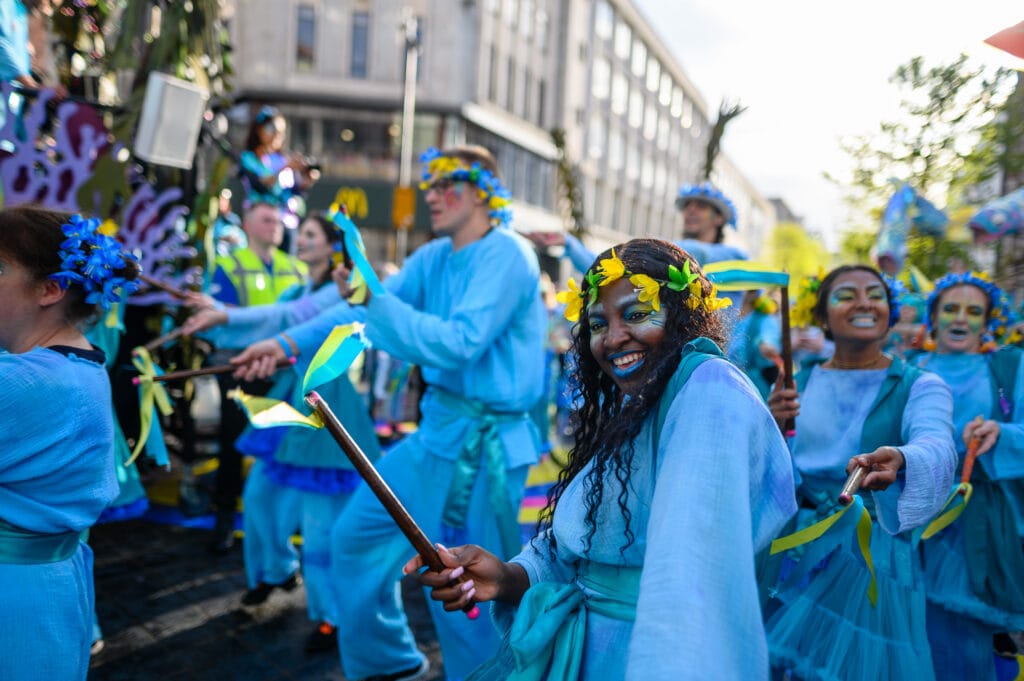 A group of performers in blue costumes with floral headbands participate in a parade. They are dancing and holding props, with some smiling at the camera. The background features a city street with buildings and spectators.