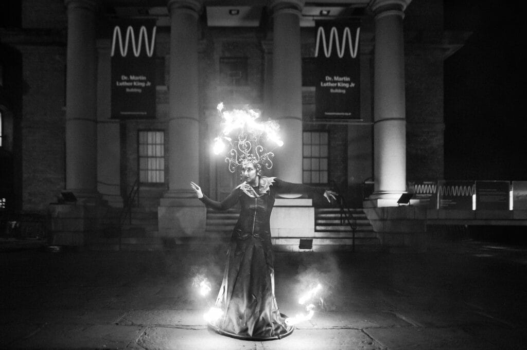 A performer in a dramatic black gown poses with fire lit around her, showcasing a fiery headpiece, against the backdrop of a historic building at night.