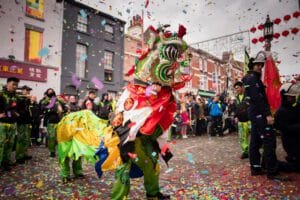 A vibrant street celebration featuring a traditional lion dance. The performer wears an elaborate lion costume in red, green, and gold, while confetti falls all around. The crowd, dressed in colorful outfits, watches the performance against a backdrop of buildings decorated with lanterns and banners.