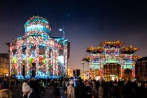 A vibrant nighttime scene featuring a historic building and an archway adorned with colorful floral projections. A crowd of people gathers in front of these illuminated structures, with traffic lights visible in the background.