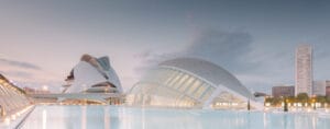 A panoramic view of the modern architectural structures designed by Santiago Calatrava in Valencia, featuring flowing lines, pointed shapes, and large glass surfaces, with a reflective body of water and a twilight sky in the background.
