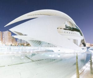 A futuristic white building designed by Calatrava, featuring sweeping curves and sharp angles, situated near a calm body of water under a twilight sky, with urban developments in the background.