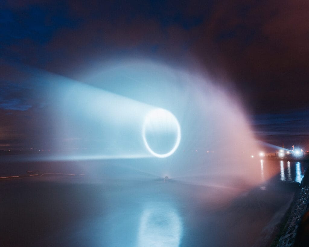 A dramatic light installation creating a circular beam of blue light against a dark, cloudy sky, reflected on the calm waters of a river in West Kirby. The setting is illuminated with soft lights from the shoreline, highlighting the atmosphere of the event.