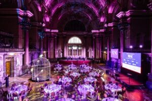 An expansive indoor venue set up for an event, featuring several round tables withtableware and centerpieces. A large decorative birdcage is positioned centrally, and a raised stage with a group of people is in the background. The venue is illuminated with purple lighting, highlighting the architecture, including tall columns and an ornate ceiling. Two large screens display the word "RISE" along with logos and information.
