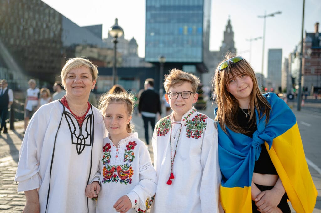 A group of four people standing outdoors. On the left, a woman wearing a traditional embroidered blouse smiles. Next to her is a young girl also in an embroidered blouse. A boy in a similar blouse stands in the center, wearing glasses. On the right, a girl draped in a blue and yellow flag poses. The background features modern buildings and streetlights.