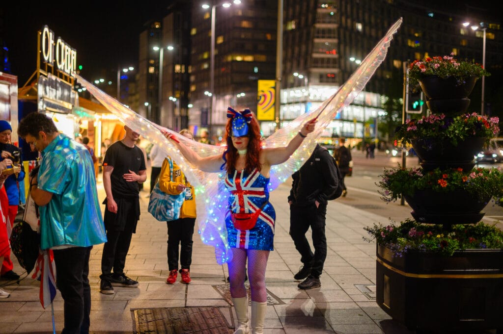 A woman dressed in a costume with Union Jack elements and illuminated wings stands in a city square at night, surrounded by people in casual clothing and a floral planter.