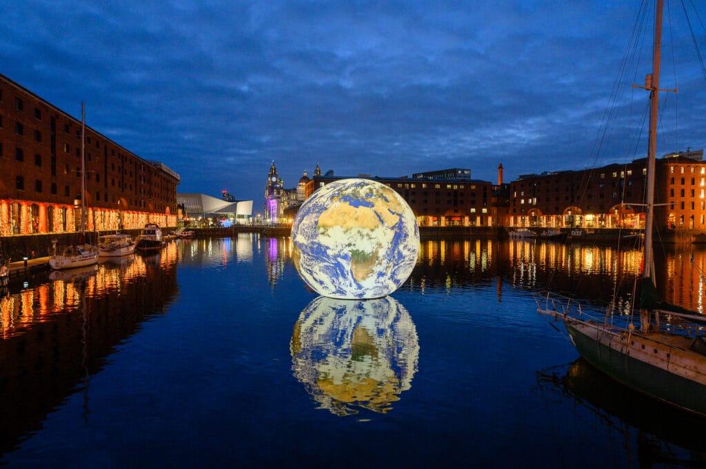 A large, illuminated globe depicting the Earth floats on the water at a dockside location, with buildings and lights reflecting in the water. The scene is set during twilight, with a cloudy sky above.