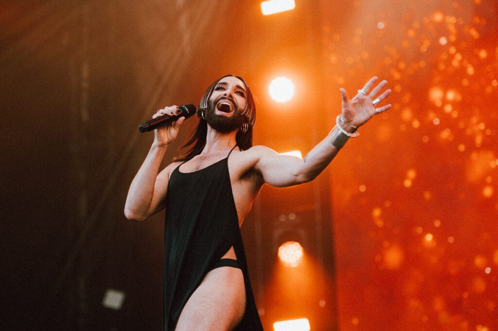 A performer with long hair and a beard wearing a sleeveless black outfit is singing into a microphone on stage, surrounded by vibrant lights in the background.