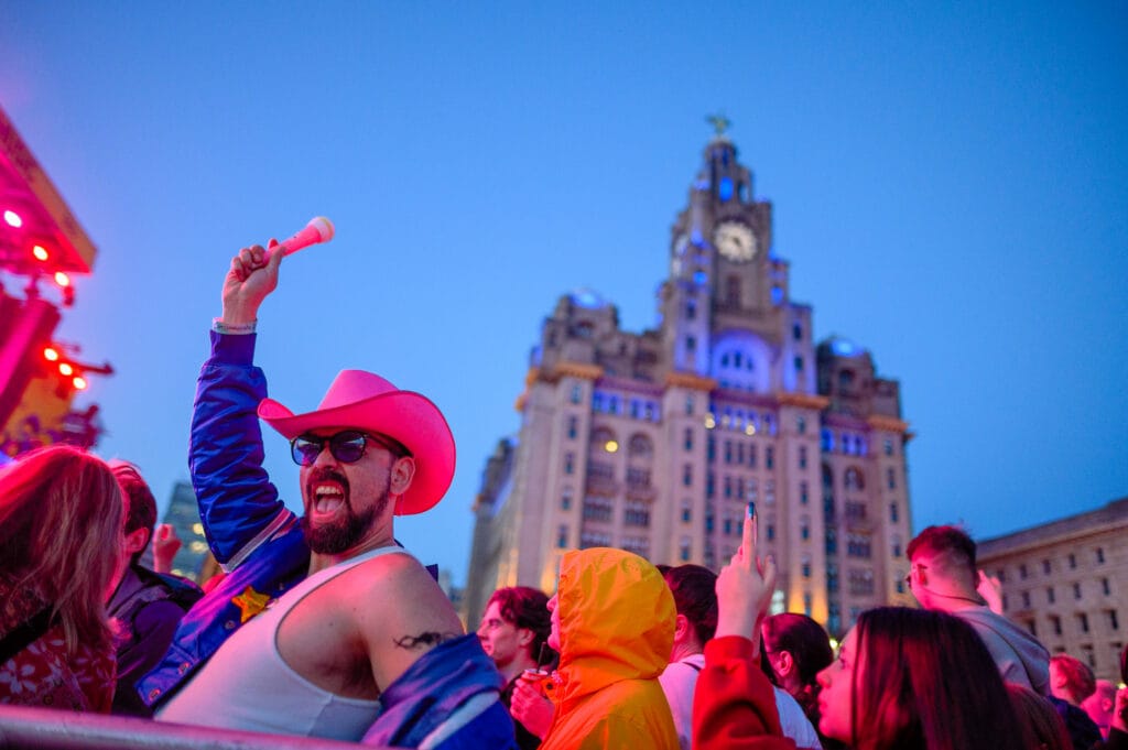 A lively outdoor scene at a festival with a crowd of people. A man wearing a pink cowboy hat and sunglasses raises a small white percussion instrument enthusiastically. The background features a prominent clock tower building illuminated against a twilight sky, with colorful stage lights visible.