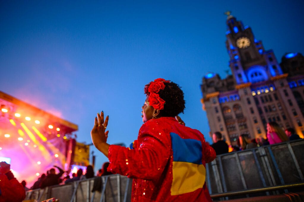 A woman in a sequined red jacket, adorned with a blue and yellow flag, dances at an outdoor concert in Liverpool, with the iconic Liver Building lit up in the background during evening twilight.