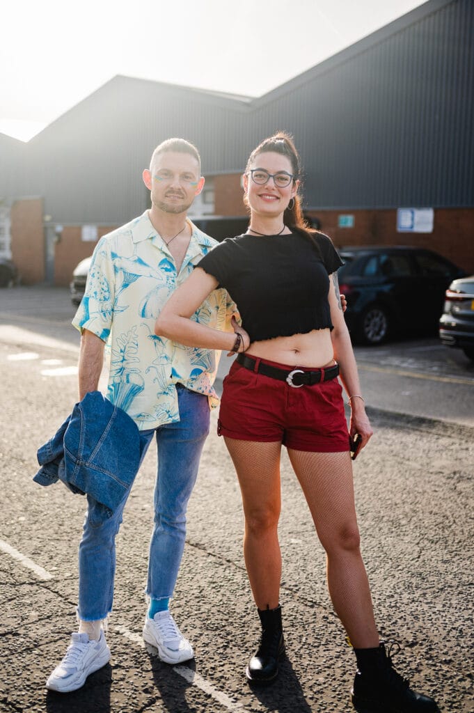 A man and a woman stand together outdoors. The man wears a light blue patterned shirt, light jeans, and white sneakers, holding a denim jacket. The woman wears a black crop top, red shorts, and black boots, with fishnet stockings. They both smile at the camera, with buildings and parked cars visible in the background.