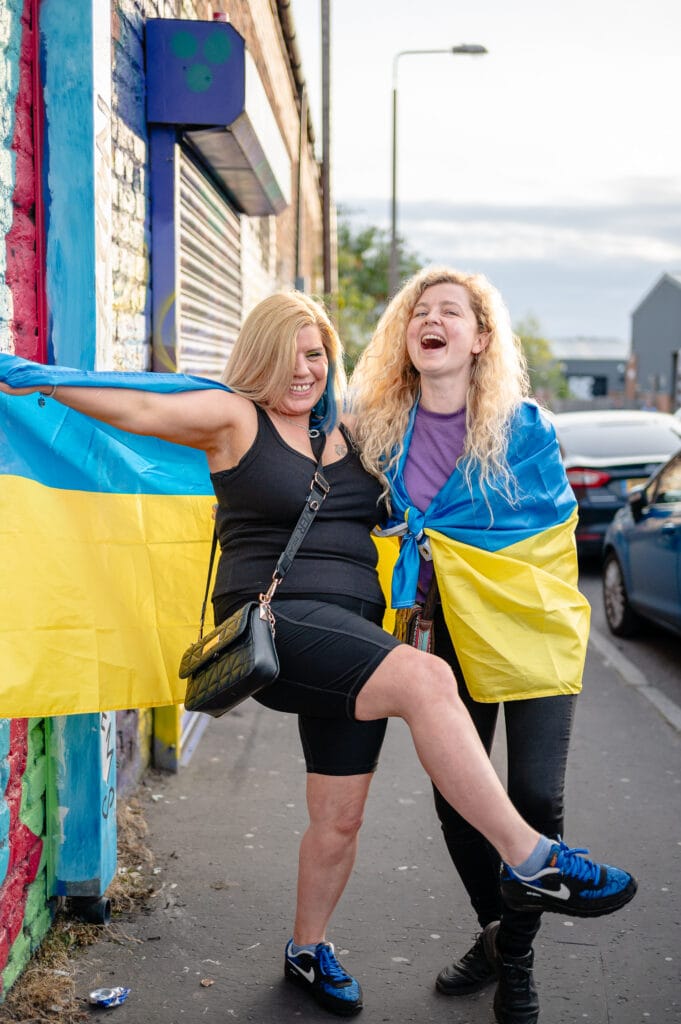 Two women standing on a street while joyfully displaying a Ukrainian flag. One woman is wearing a black dress and has long blonde hair, and the other has curly blonde hair and is dressed in a light shirt and dark pants. They are posing playfully, with one woman lifting her leg.