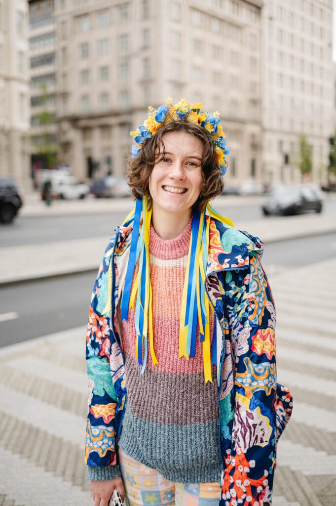A young woman with curly hair is smiling at the camera while wearing a colorful sweater and an ornate flower crown with blue and yellow ribbons. She is standing outdoors with buildings and cars in the background.