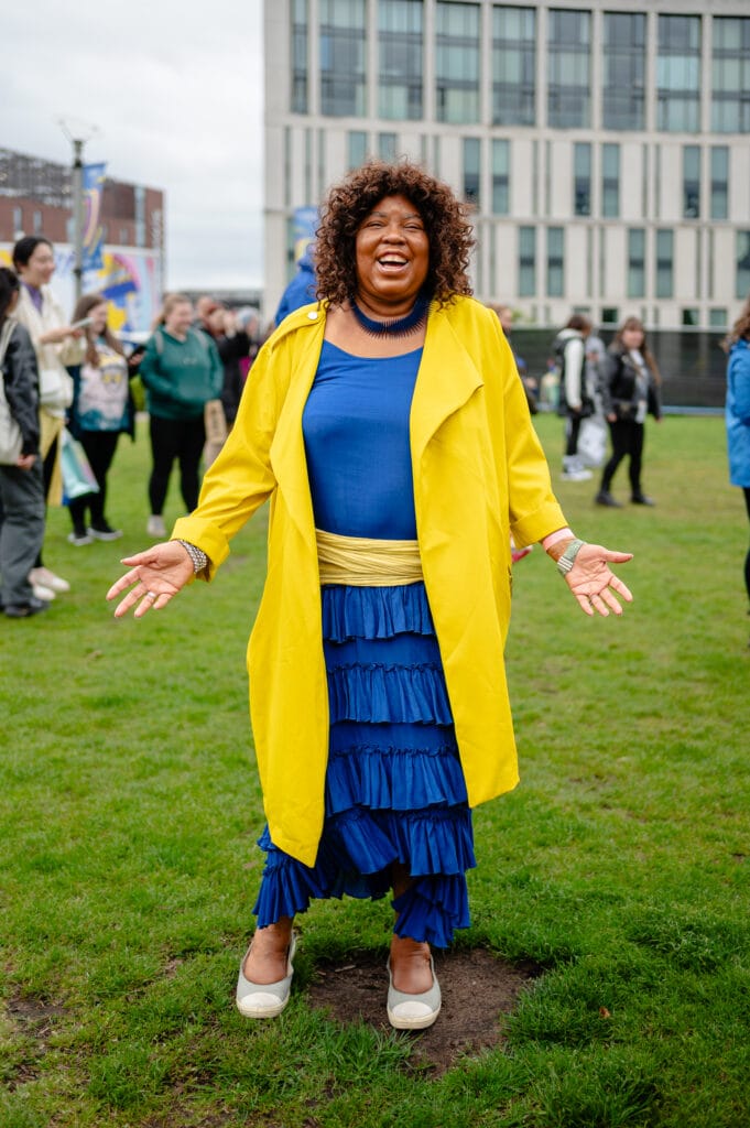 A person stands on a grass field with their arms outstretched, wearing a bright yellow coat over a blue top and a tiered blue skirt. They have curly hair and are smiling widely. In the background, a crowd of people are visible along with a modern building.