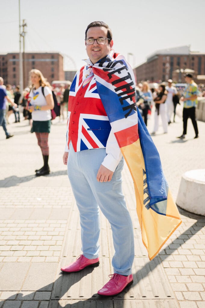 A man stands outdoors wearing a bright outfit consisting of a vest with the British flag and light blue trousers. He has a multicolored flag draped over his shoulders and is smiling. The background shows other people and buildings in a sunny setting.