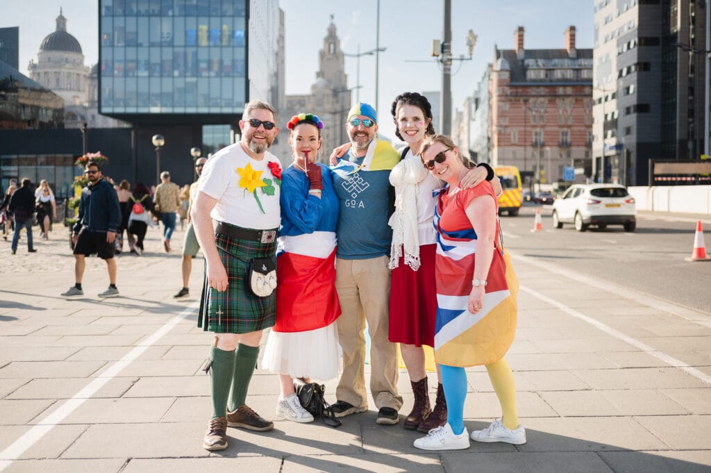 A group of five people dressed in colorful costumes posing together on a street. They include a man in a kilt with a t-shirt featuring a flower design, a woman in a blue top and a red skirt, another person wearing a blue and yellow headband, and two women dressed in outfits with British flag patterns. The background features a mix of modern and historical buildings.
