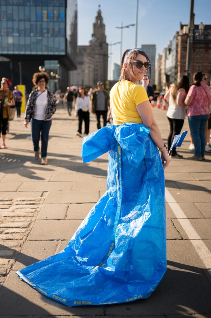 A woman wearing a bright yellow top and sunglasses stands in a busy urban area. She is dressed in a large blue garment made from what appears to be a plastic IKEA bag, which trails behind her on the ground. Several other people are seen walking in the background.