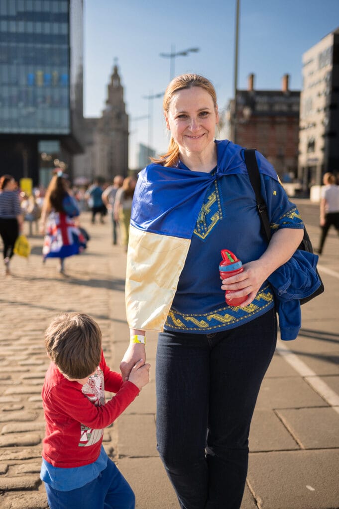 A woman wearing a blue and yellow garment, resembling the Ukrainian flag, stands on a city street holding hands with a child dressed in red. The background features various buildings and people walking.